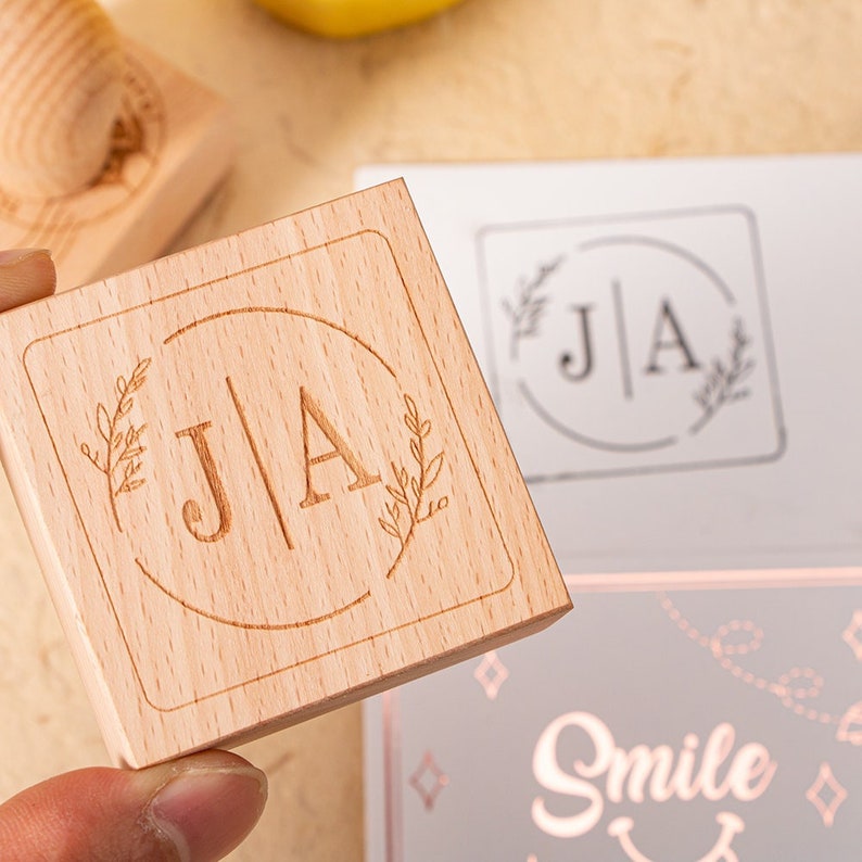 May include: A wooden stamp with the initials "J|A" in a circle surrounded by small branches. The stamp is on a light brown surface.