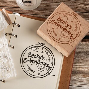 May include: A wooden stamp with a circular design featuring the text "Becky's Embroideries" surrounded by a floral pattern, a butterfly, and a needle and thread. The stamp is on a notebook with a white lace doily.