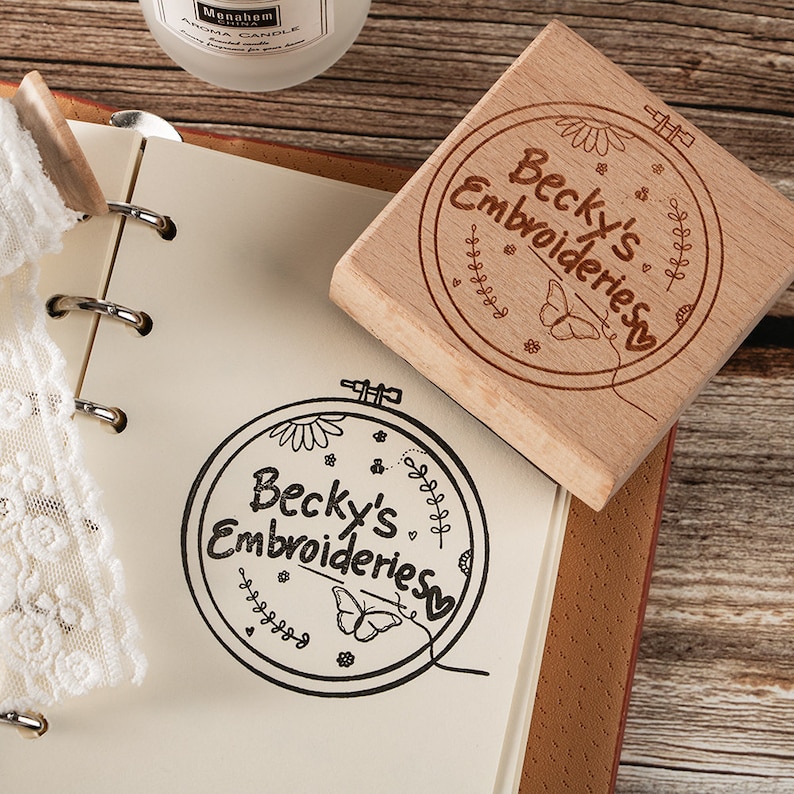 May include: A wooden stamp with a circular design featuring the text "Becky's Embroideries" surrounded by a floral pattern, a butterfly, and a needle and thread. The stamp is on a notebook with a white lace doily.