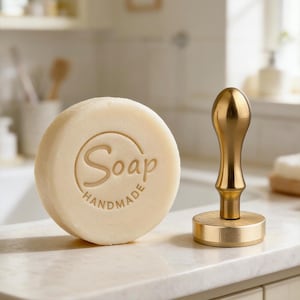 May include: A round, light-colored handmade soap with the word "Soap" and "Handmade" engraved on its surface. A gold-colored metal stamp is next to the soap. The soap and stamp are on a white countertop.