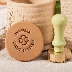 Puede incluir: Un sello de cerámica marrón claro con el texto "CHAFFERS LOUISE'S POTTERY" que rodea un diseño de trébol de cuatro hojas. Un mango de sello verde claro con una base dorada está al lado del sello de cerámica. El fondo incluye una maceta de cerámica.