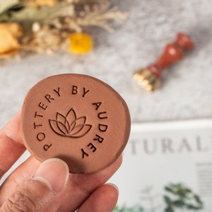 Könnte beinhalten: Ein runder, terrakottafarbener Töpferstempel mit dem Text "POTTERY BY AUDREY", der ein Lotusblumen-Design umrandet. Im Hintergrund ist ein verschwommenes Wachssiegel zu sehen. Der Stempel wird in einer Hand gehalten.