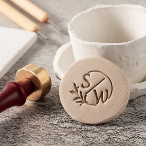 Puede incluir: Un sello redondo de color claro con las iniciales "SW" y un diseño de hojas. Un sello dorado con un mango rojo oscuro está en primer plano. Una taza de cerámica blanca y otras herramientas están en el fondo.