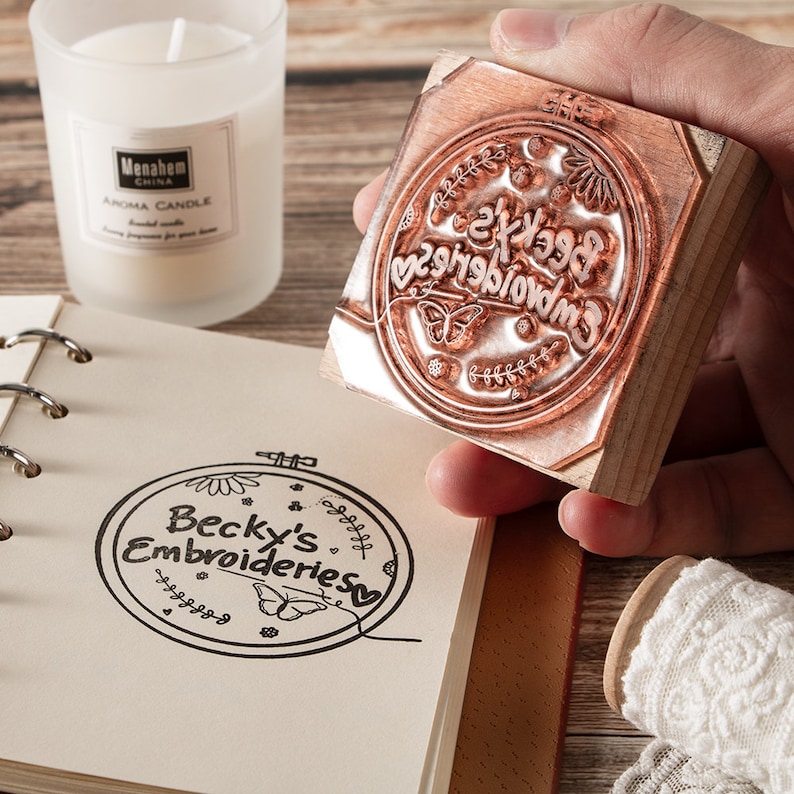 May include: Wooden stamp with a circular design featuring the text "Becky's Embroideries" surrounded by floral and butterfly motifs. The stamp is being held in a hand.