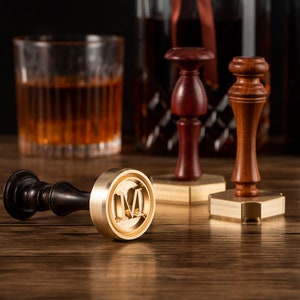 Könnte beinhalten: Drei Wachssiegelstempel aus Holz mit Messingstempelköpfen, einer mit dem Buchstaben "M". Die Stempel haben dunkelbraune Holzgriffe und werden auf einer Holzoberfläche präsentiert. Ein Glas bernsteinfarbener Flüssigkeit und eine Flasche befinden sich im Hintergrund.
