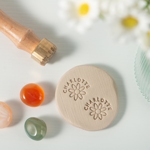 Puede incluir: Un sello de madera con una cabeza de latón, con el nombre "Charlotte" y un diseño de margarita. El sello se utiliza para crear impresiones en una superficie de arcilla marrón claro.