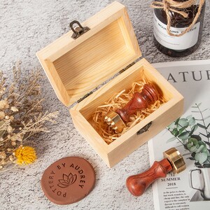 Pode incluir: Uma caixa de madeira com tampa articulada, contendo um selo de cera vermelho e dourado aninhado em palha. Um segundo selo e um porta-copos de terracota com o texto "Pottery by Audrey" também são visíveis.