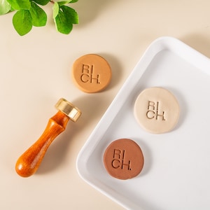 May include: Three round clay stamps with the word "RICH" embossed on them. The stamps are in shades of brown, beige, and terracotta. A wooden handle with a brass top is also included.