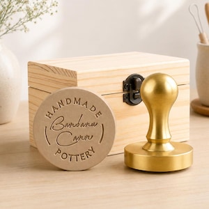 May include: A brass pottery stamp with a rounded handle and a circular base, accompanied by a circular clay impression featuring the text "HANDMADE Barbara Cann POTTERY" and a light-coloured wooden storage box with a metal latch closure.