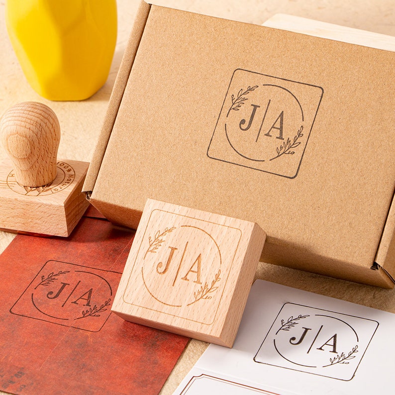 May include: A set of two wooden stamps with a floral design and the initials "J|A" in a circle. The stamps are packaged in a brown cardboard box.