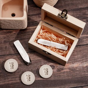 Könnte beinhalten: Ein Satz von drei Metallstempeln mit einem Sonnendesign und dem Text "JaRa 999". Die Stempel befinden sich in einer Holzkiste mit einem Scharnierdeckel und einem Metallverschluss. Die Kiste ist mit zerkleinertem Papier gefüllt.