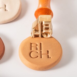 May include: A brass stamp with the word "RICH" on it, used to create impressions on clay or other materials. The stamp is attached to a wooden handle.