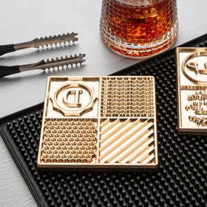 May include: A gold-coloured square coaster featuring four different textured patterns. It rests on a black bar mat, beside a rocks glass filled with amber liquid and a pair of ice tongs.