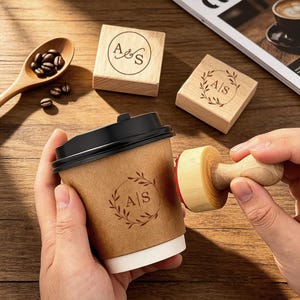 Könnte beinhalten: Ein brauner Pappbecher mit schwarzem Deckel wird mit einem Holzstempel gestempelt. Der Stempel und der Becher zeigen ein kreisförmiges Design mit den Buchstaben "A" und "S" in einem Kranz. Kaffeebohnen und Holzstempel sind ebenfalls sichtbar.