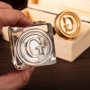 Puede incluir: Un cubito de hielo transparente y cuadrado con la letra "G" en relieve en un diseño circular. Un sello de metal dorado con la letra "G" es visible en el fondo, junto con una caja de madera. El cubito de hielo es sostenido por una mano.