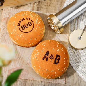 May include: Two sesame seed hamburger buns with stamped text. One bun reads "MADE BY BOB" in a circle. The other bun has the letters "A & B" with a leaf symbol. A branding iron is in the background.