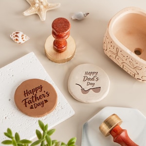 Könnte beinhalten: Eine Sammlung von Vatertagsgeschenken, darunter ein Holzstempel mit Messingfuß, Untersetzer mit den Texten "Happy Father's Day" und "Happy Dad's Day" sowie dekorative Muscheln. Der Stempel hat einen dunklen Holzgriff.