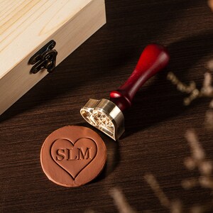 Op de afbeelding: Een hartvormige messing stempel met een houten handvat, gebruikt om waszegels te maken. De stempel heeft de initialen "SLM" in een hart. Een hartvormige zegel van rode klei met dezelfde initialen is ook te zien.