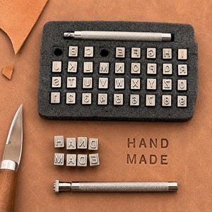 Puede incluir: Un set de marroquinería que incluye una herramienta con mango de metal, un organizador de espuma rectangular con sellos metálicos plateados de letras y números, y sellos adicionales que forman las palabras HAND MADE en el cuero.