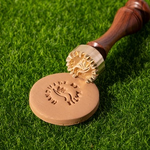 May include: A brass stamp with a wooden handle, featuring a design of a hand holding a leaf. The stamp is being used to create an impression on a brown clay surface. The impression reads "Handmade by"