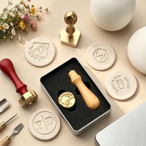 May include: A wax seal stamp kit featuring a wooden handle, a brass stamp head, and a metal storage tin. Included are several circular clay discs embossed with various custom designs, including initials and dates, alongside metal sculpting tools.