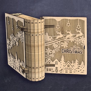 Peut inclure: D&eacute;coration en bois en forme de livre sur le th&egrave;me de No&euml;l. Le livre ouvert r&eacute;v&egrave;le un motif d&eacute;coup&eacute; au laser d'un village d'hiver, de flocons de neige, d'&eacute;toiles et du tra&icirc;neau du P&egrave;re No&euml;l. Les mots "Merry Christmas" sont &eacute;galement visibles.