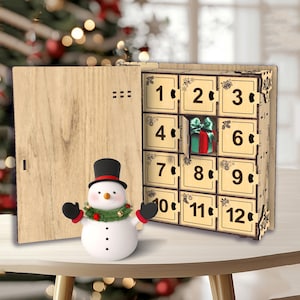Peut inclure: Un calendrier de l'Avent en bois avec des compartiments num&eacute;rot&eacute;s, dont un ouvert pour r&eacute;v&eacute;ler un petit cadeau. Un bonhomme de neige avec un chapeau haut de forme noir et une guirlande verte se tient devant le calendrier.