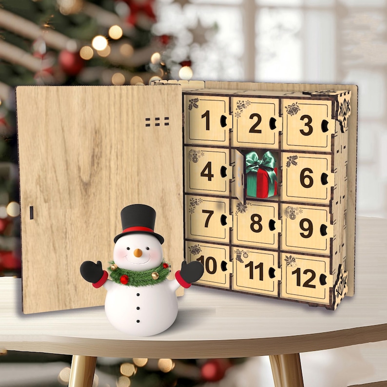 Peut inclure: Un calendrier de l'Avent en bois avec des compartiments num&eacute;rot&eacute;s, dont un ouvert pour r&eacute;v&eacute;ler un petit cadeau. Un bonhomme de neige avec un chapeau haut de forme noir et une guirlande verte se tient devant le calendrier.