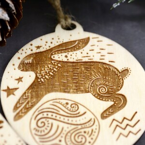 May include: A wooden ornament featuring a stylized rabbit with intricate details, surrounded by stars and a swirling pattern. The ornament is a circular shape and has a string for hanging.