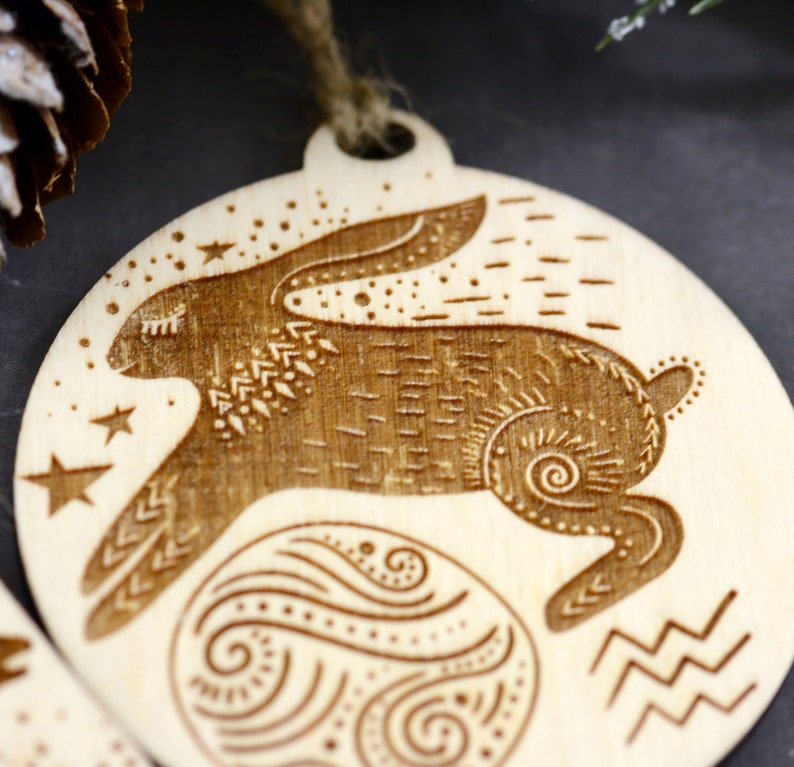 May include: A wooden ornament featuring a stylized rabbit with intricate details, surrounded by stars and a swirling pattern. The ornament is a circular shape and has a string for hanging.