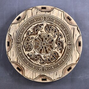 K&ouml;nnte beinhalten: Eine runde Holzplakette mit einem geschnitzten Design eines Kompasses mit vier Punkten, umgeben von einem Knotenmuster und vier mythischen Kreaturen. Die Plakette hat eine hellbraune Holzmaserung und ist mit einer dunkelbraunen Schnitzerei verziert.