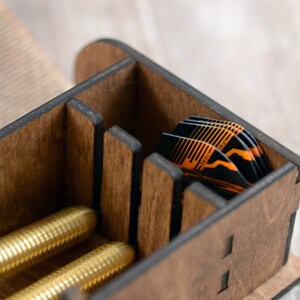 May include: A wooden box with multiple compartments for storing darts. The box is made of dark wood with black accents. The box is open and you can see a stack of orange and black darts inside.