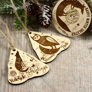 May include: Three wooden ornaments with a winter solstice theme. The ornaments are shaped like triangles and feature a wolf, an owl, and a fox. The ornaments are decorated with stars, moons, and other celestial symbols. The text on the ornaments reads "Yuletide Greetings", "Blessed Yule", and "Happy Winter Solstice".