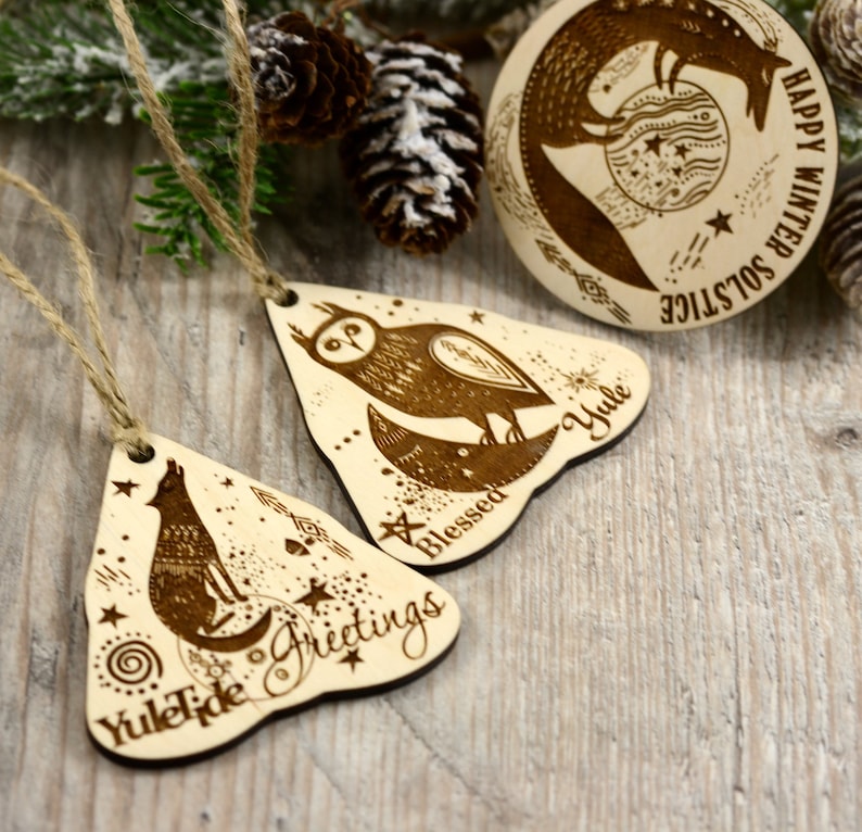 May include: Three wooden ornaments with a winter solstice theme. The ornaments are shaped like triangles and feature a wolf, an owl, and a fox. The ornaments are decorated with stars, moons, and other celestial symbols. The text on the ornaments reads "Yuletide Greetings", "Blessed Yule", and "Happy Winter Solstice".