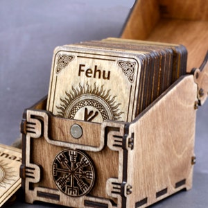 May include: Wooden box with a hinged lid, containing a set of wooden rune cards. The top card features the rune "Fehu" and a sun symbol. The bottom of the box has a compass design.