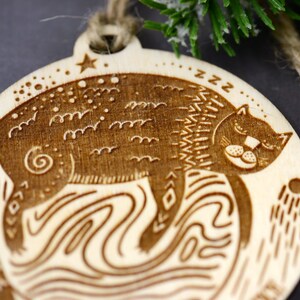 May include: A wooden Christmas ornament featuring a stylized cat sleeping on a wave pattern. The ornament is brown and has a star and the word "zzz" engraved on it.