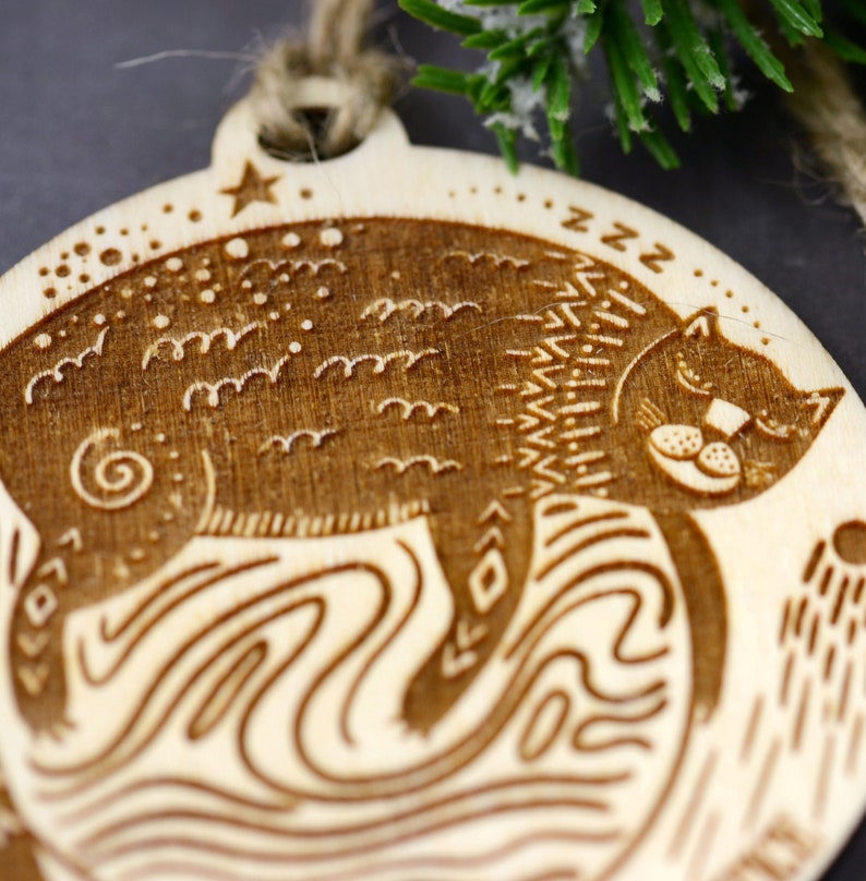 May include: A wooden Christmas ornament featuring a stylized cat sleeping on a wave pattern. The ornament is brown and has a star and the word "zzz" engraved on it.
