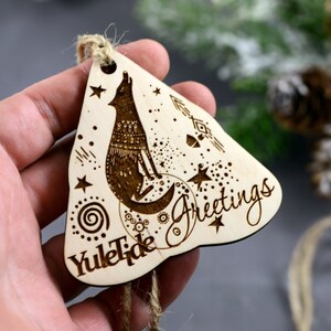 May include: Wooden Christmas ornament with a wolf howling at the moon, surrounded by stars and a spiral design. The ornament is inscribed with "Yuletide Greetings".