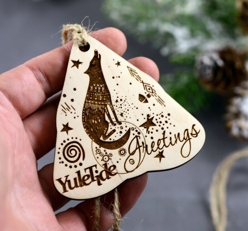 May include: Wooden Christmas ornament with a wolf howling at the moon, surrounded by stars and a spiral design. The ornament is inscribed with "Yuletide Greetings".