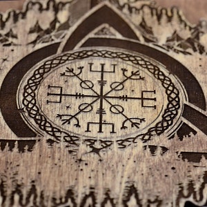 May include: A wooden sign with a black and white compass design surrounded by a Celtic knot. The compass is a Vegvisir, a Norse symbol for guidance and protection. The background is a wood grain pattern with a forest scene.