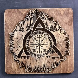 May include: A wooden wall hanging with a brown stain. The design is a black and white compass with a Celtic knot design around it. The compass is surrounded by a triangle with a mountain and trees in the background.