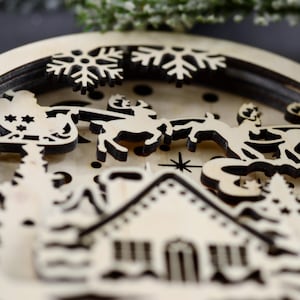 Pu&ograve; includere: Un ornamento natalizio in legno con un design a strati che presenta una casa, alberi, fiocchi di neve e una renna che tira una slitta. L'ornamento &egrave; realizzato in legno chiaro con accenti marrone scuro.