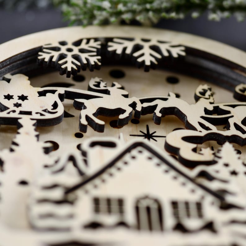 Pu&ograve; includere: Un ornamento natalizio in legno con un design a strati che presenta una casa, alberi, fiocchi di neve e una renna che tira una slitta. L'ornamento &egrave; realizzato in legno chiaro con accenti marrone scuro.