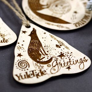 May include: Wooden Christmas ornament with a wolf howling at the moon, surrounded by stars and a spiral design. The ornament is shaped like a teardrop and has the text "Yuletide Greetings" on it.