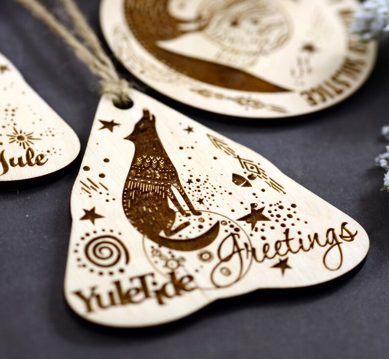 May include: Wooden Christmas ornament with a wolf howling at the moon, surrounded by stars and a spiral design. The ornament is shaped like a teardrop and has the text "Yuletide Greetings" on it.