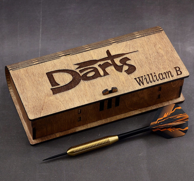 May include: A wooden box with the word "Darts" engraved on the top. The box is personalized with the name "William B". A black and orange dart is lying next to the box.