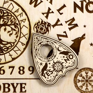 K&ouml;nnte beinhalten: Eine Holzplanchette mit einem schwarz-wei&szlig;en Design, das einen Halbmond, Sterne, eine Sonne mit einem Kompass und zwei Raben zeigt. Die Planchette tr&auml;gt die Worte "YES" und "NO" ein. Die Planchette liegt auf einem Ouija-Brett aus Holz mit einem Lebensbaumdesign, Zahlen und Buchstaben.