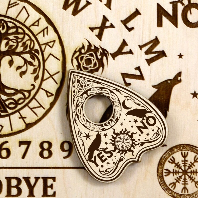K&ouml;nnte beinhalten: Eine Holzplanchette mit einem schwarz-wei&szlig;en Design, das einen Halbmond, Sterne, eine Sonne mit einem Kompass und zwei Raben zeigt. Die Planchette tr&auml;gt die Worte "YES" und "NO" ein. Die Planchette liegt auf einem Ouija-Brett aus Holz mit einem Lebensbaumdesign, Zahlen und Buchstaben.