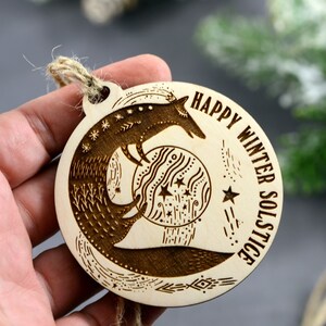 May include: A wooden ornament with a laser-engraved design of a fox holding a moon with stars. The text "Happy Winter Solstice" is engraved around the edge of the ornament.