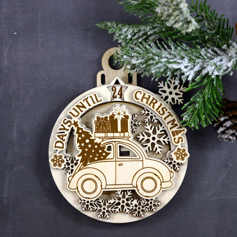 Puede incluir: Un adorno de cuenta atr&aacute;s para Navidad de madera con forma de c&iacute;rculo con un coche que lleva un &aacute;rbol de Navidad y regalos. El adorno tiene el texto "Days Until Christmas" y el n&uacute;mero "24".